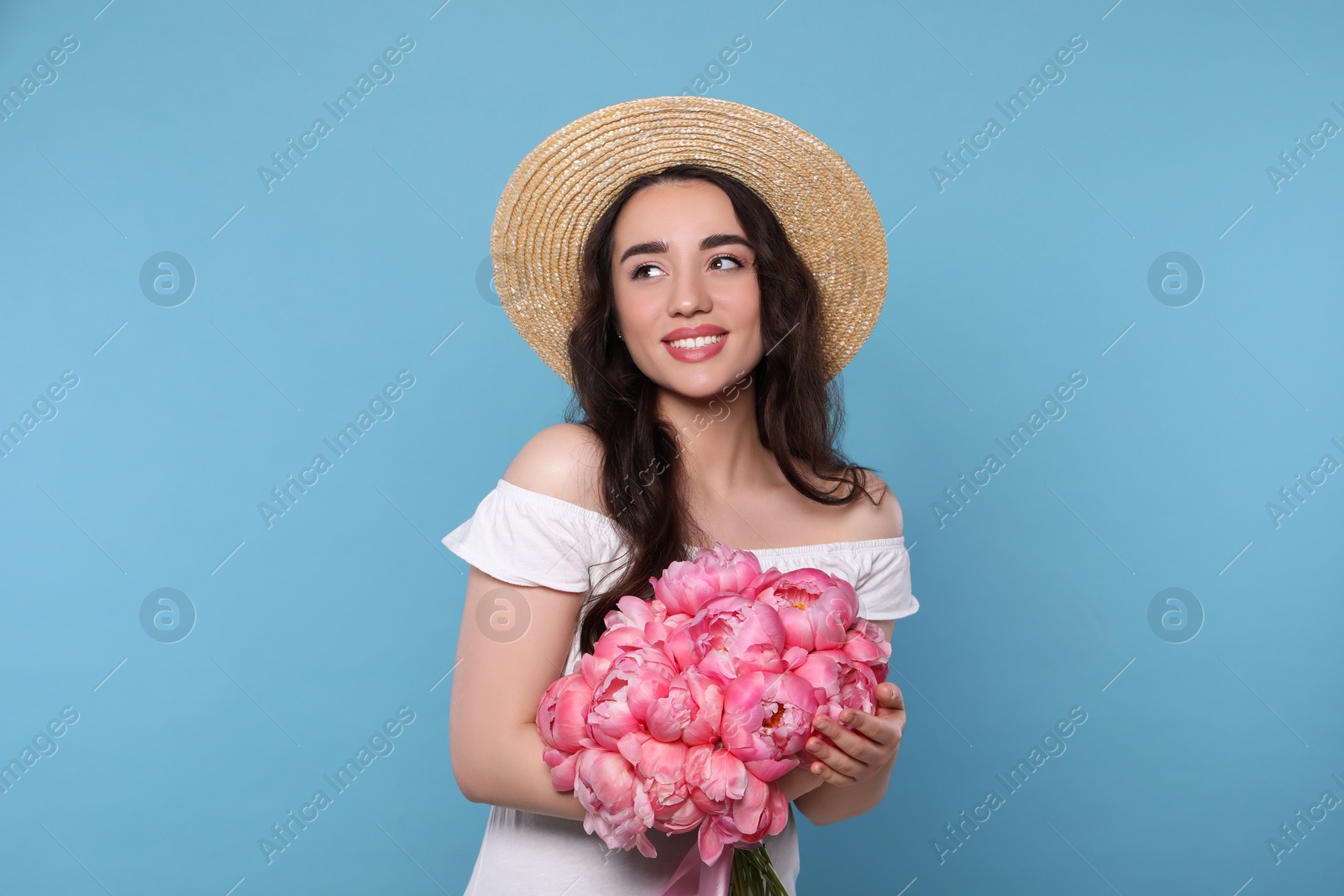 Photo of Beautiful young woman in straw hat with bouquet of pink peonies against light blue background