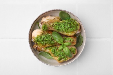 Delicious fried chicken drumsticks with pesto sauce and basil in bowl on white tiled table, top view