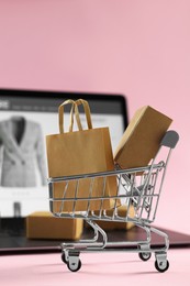 Photo of Online store. Mini shopping cart, purchases and laptop on pink background, selective focus. Space for text