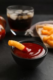 Photo of Tasty cheesy corn stick in bowl of red sauce on black table, closeup