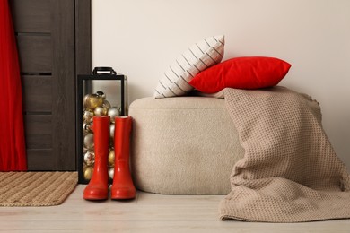 Photo of Container with baubles, ottoman and red rubber boots near wooden door in room. Christmas decoration