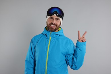 Photo of Winter sports. Happy man in ski suit and goggles showing V-sign on gray background