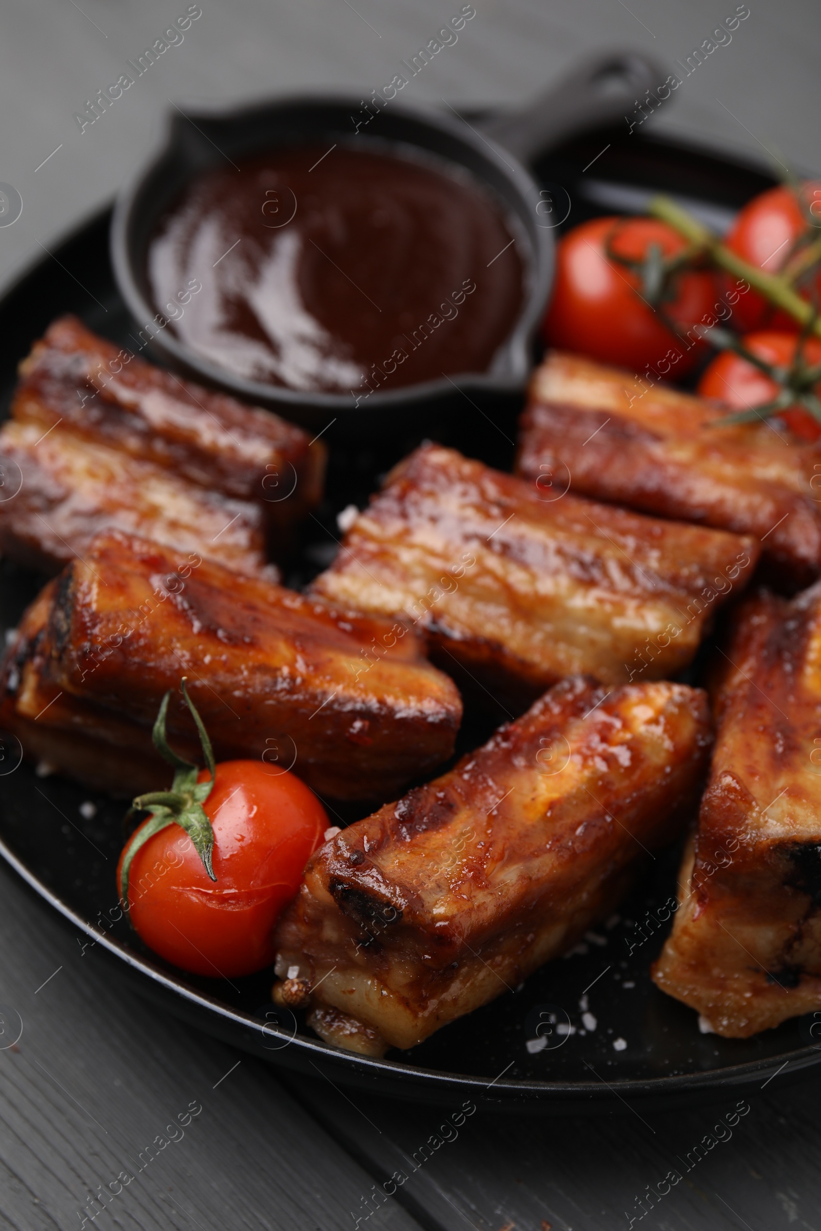 Photo of Tasty roasted pork ribs served with sauce and tomatoes on grey wooden table, closeup