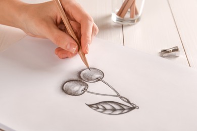 Photo of Woman drawing cherries with graphite pencil on paper at white wooden table, closeup