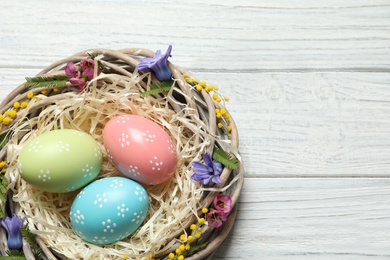 Photo of Wicker nest with painted Easter eggs and flowers on wooden table, top view. Space for text