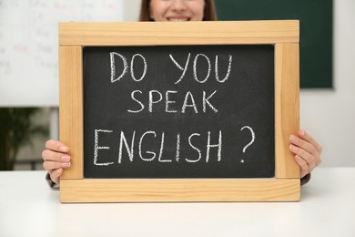 Teacher holding small chalkboard with inscription Do You Speak English? at table in classroom, closeup