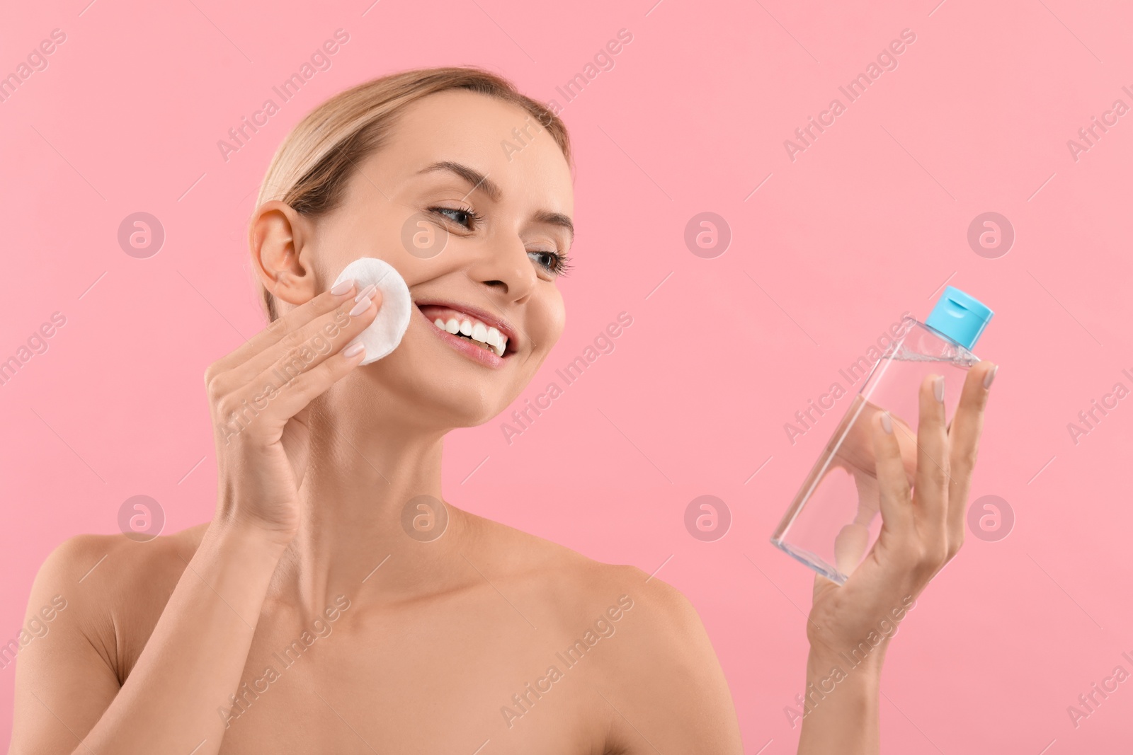 Photo of Smiling woman removing makeup with cotton pad and holding bottle on pink background