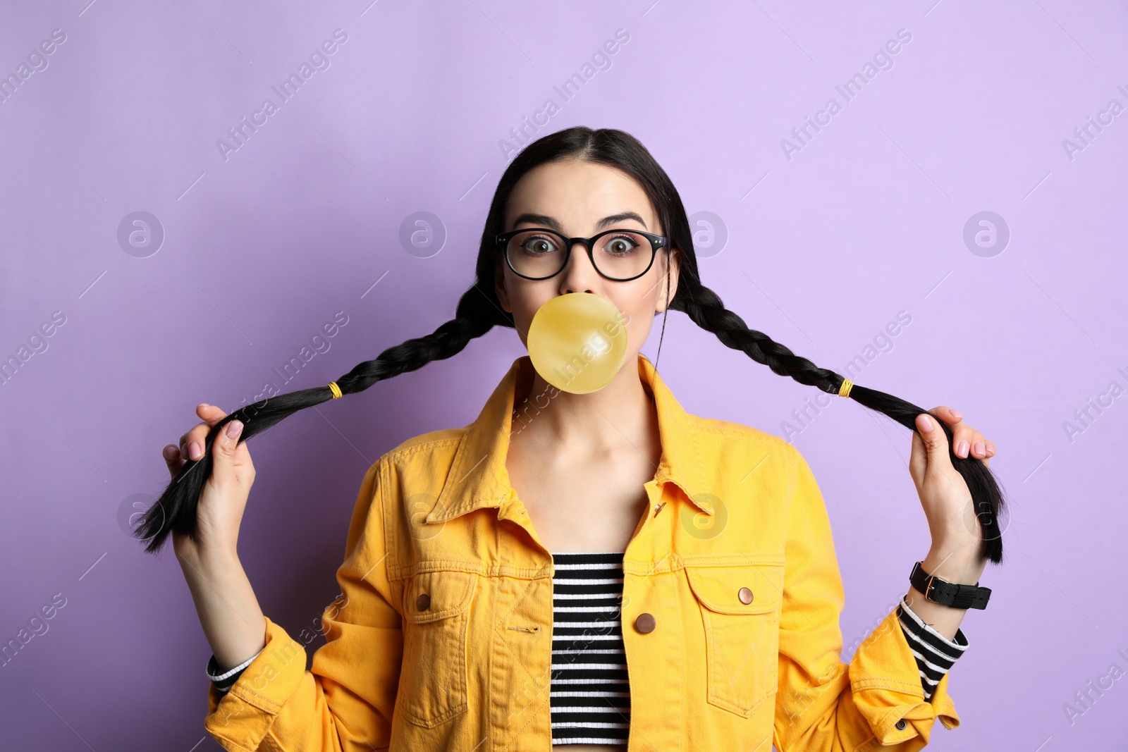 Photo of Fashionable young woman with braids blowing bubblegum on lilac background