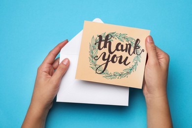 Photo of Woman holding envelope and card with phrase Thank you on light blue background, top view