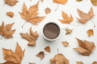 Cup of hot drink and autumn leaves on white wooden table, flat lay. Cozy atmosphere