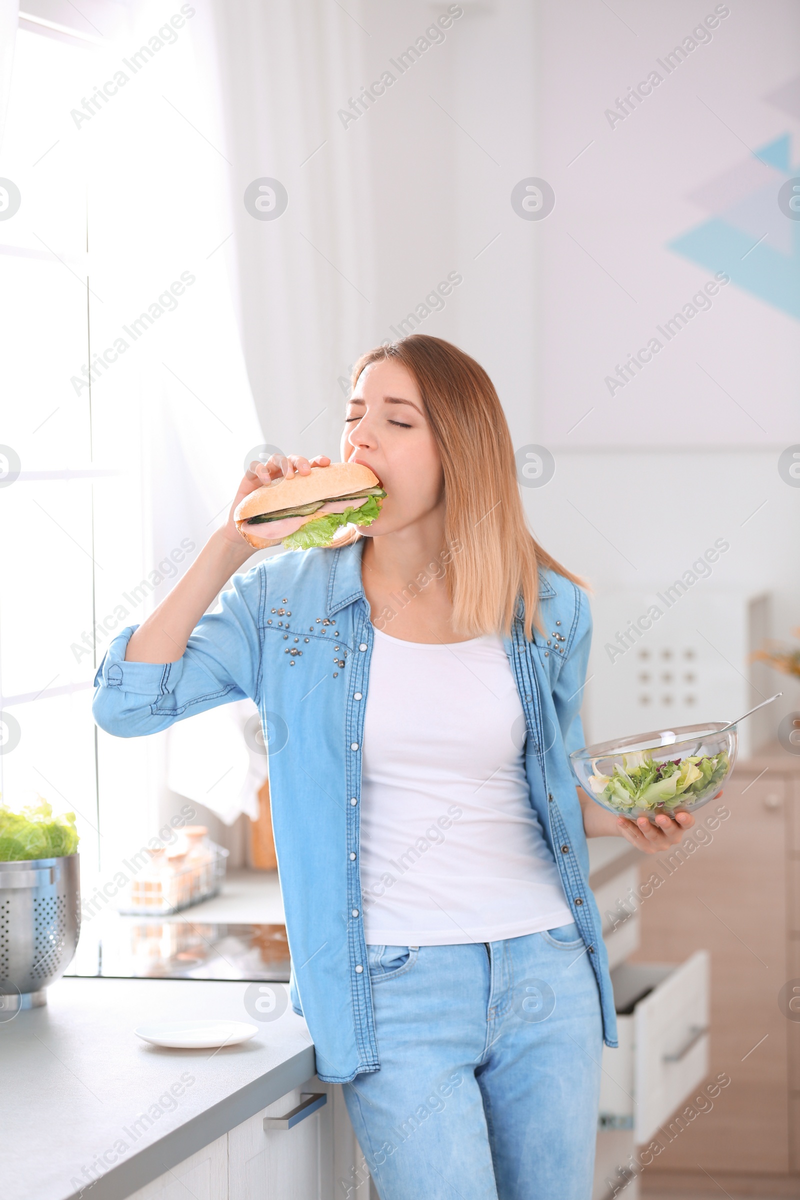 Photo of Beautiful young woman eating sandwich instead of salad in kitchen. Failed diet