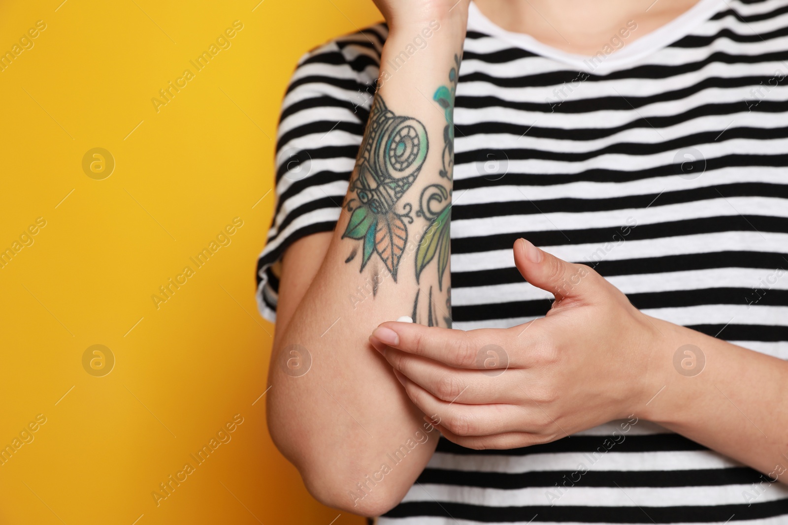 Photo of Woman applying cream on her arm with tattoos against yellow background, closeup. Space for text