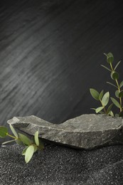 Photo of Presentation of product. Stone podium and green branches on black sand against dark textured background. Space for text