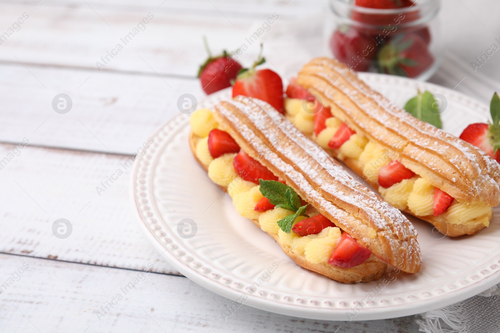 Photo of Delicious eclairs filled with cream, strawberries and mint on white wooden table, closeup