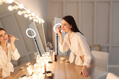 Photo of Young woman applying make up near illuminated mirror indoors