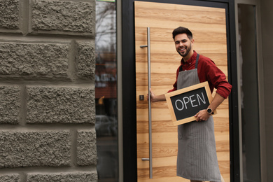Young male business owner holding OPEN sign near his cafe. Space for text