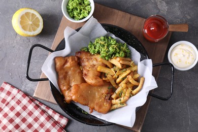 Photo of Tasty fish, chips, sauces and peas on grey table, flat lay