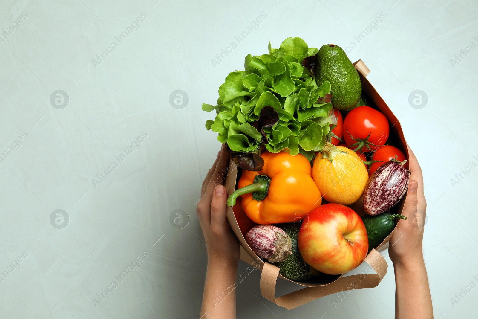Photo of Woman holding paper bag with fresh vegetables and fruits on light background, top view. Space for text
