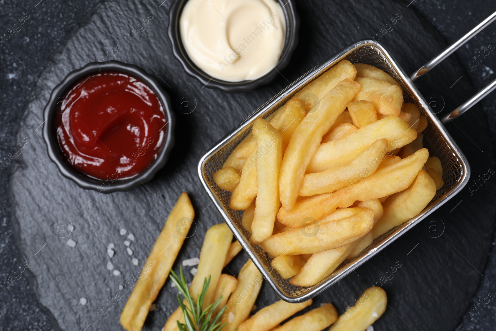 Photo of Tasty French fries, ketchup, rosemary and mayonnaise on black textured table, top view