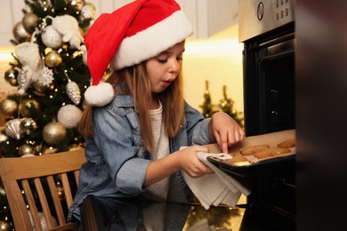 Photo of Little child in Santa hat taking baking sheet with Christmas cookies out of oven indoors