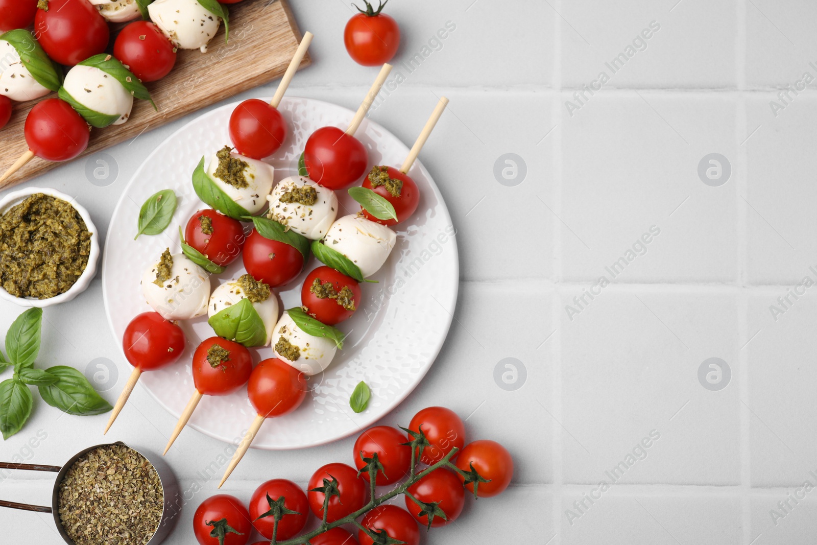 Photo of Caprese skewers with tomatoes, mozzarella balls and basil on white tiled table, flat lay. Space for text