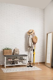 Photo of Stylish hallway interior with coat rack, shoe storage bench and mirror near white brick wall