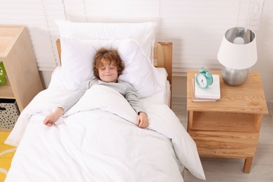 Photo of Sleepy little boy lying in cosy bed near alarm clock on bedside table indoors
