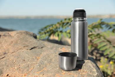 Photo of Modern silver thermos on stone near river. Space for text