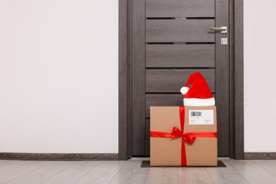 Photo of Christmas gift box and Santa hat near door indoors, space for text. Sending present by mail