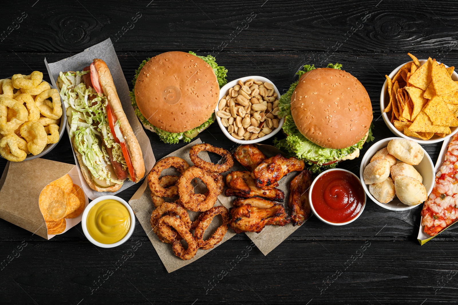 Photo of Burgers, onion rings and other fast food on black wooden table, flat lay