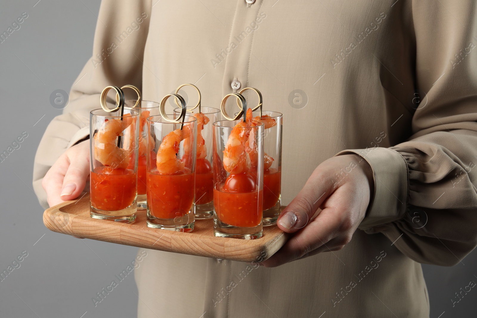 Photo of Woman holding tasty canapes with shrimps, tomatoes and sauce in shot glasses on grey background, closeup