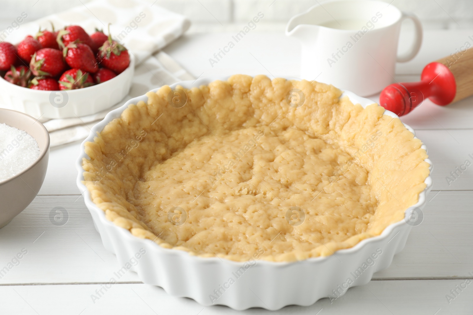 Photo of Making shortcrust pastry. Raw dough in baking dish, strawberries, sugar, rolling pin and milk on white wooden table