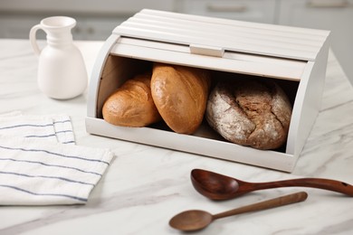 Wooden bread basket with freshly baked loaves and spoons on white marble table in kitchen