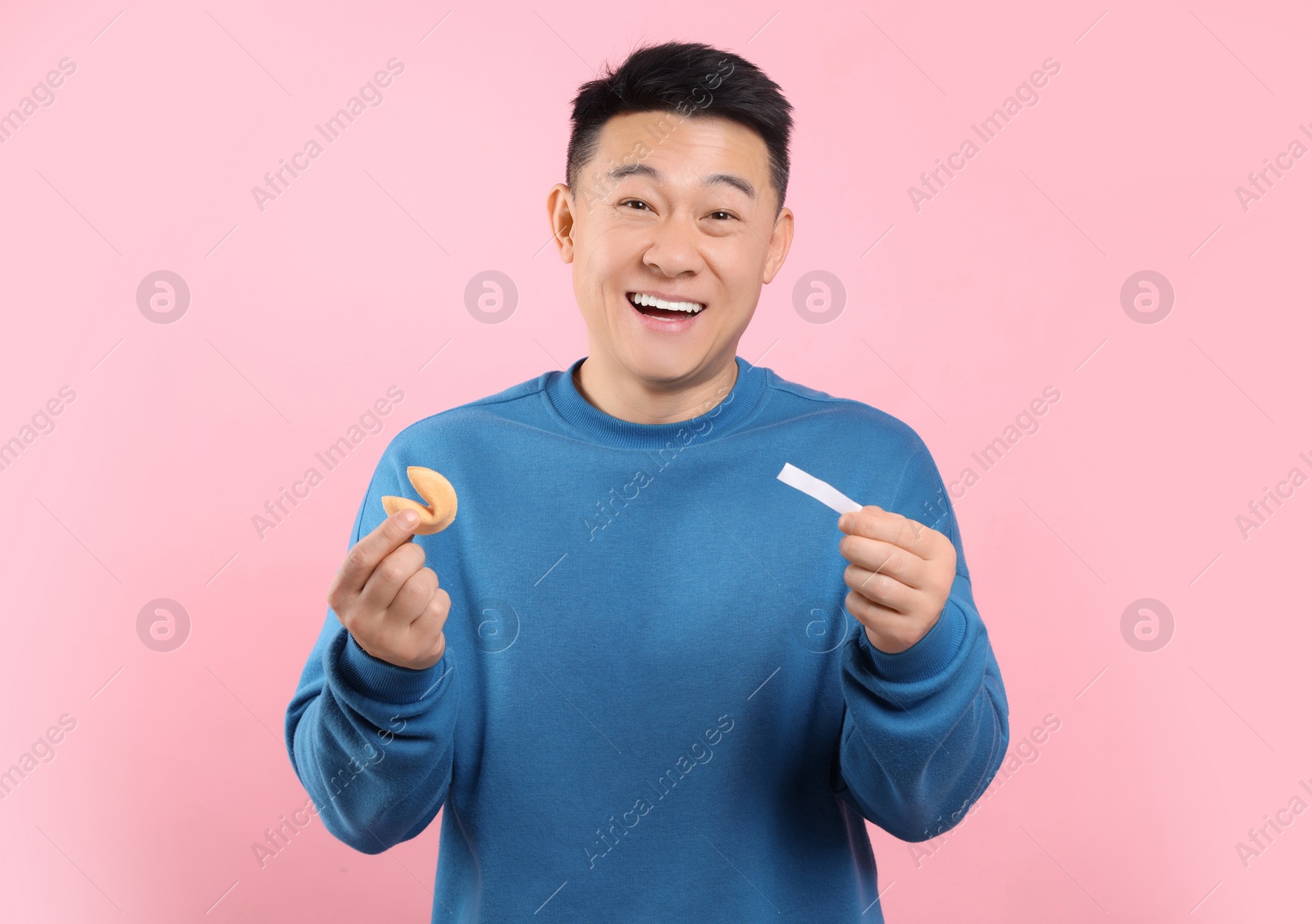 Photo of Happy asian man holding tasty fortune cookie with prediction on pink background