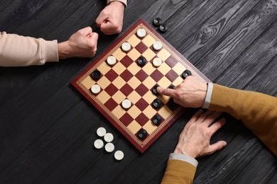 Photo of Senior man playing checkers with angry partner at black wooden table, top view
