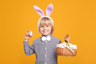 Happy boy in bunny ears headband holding wicker basket with painted Easter eggs on orange background