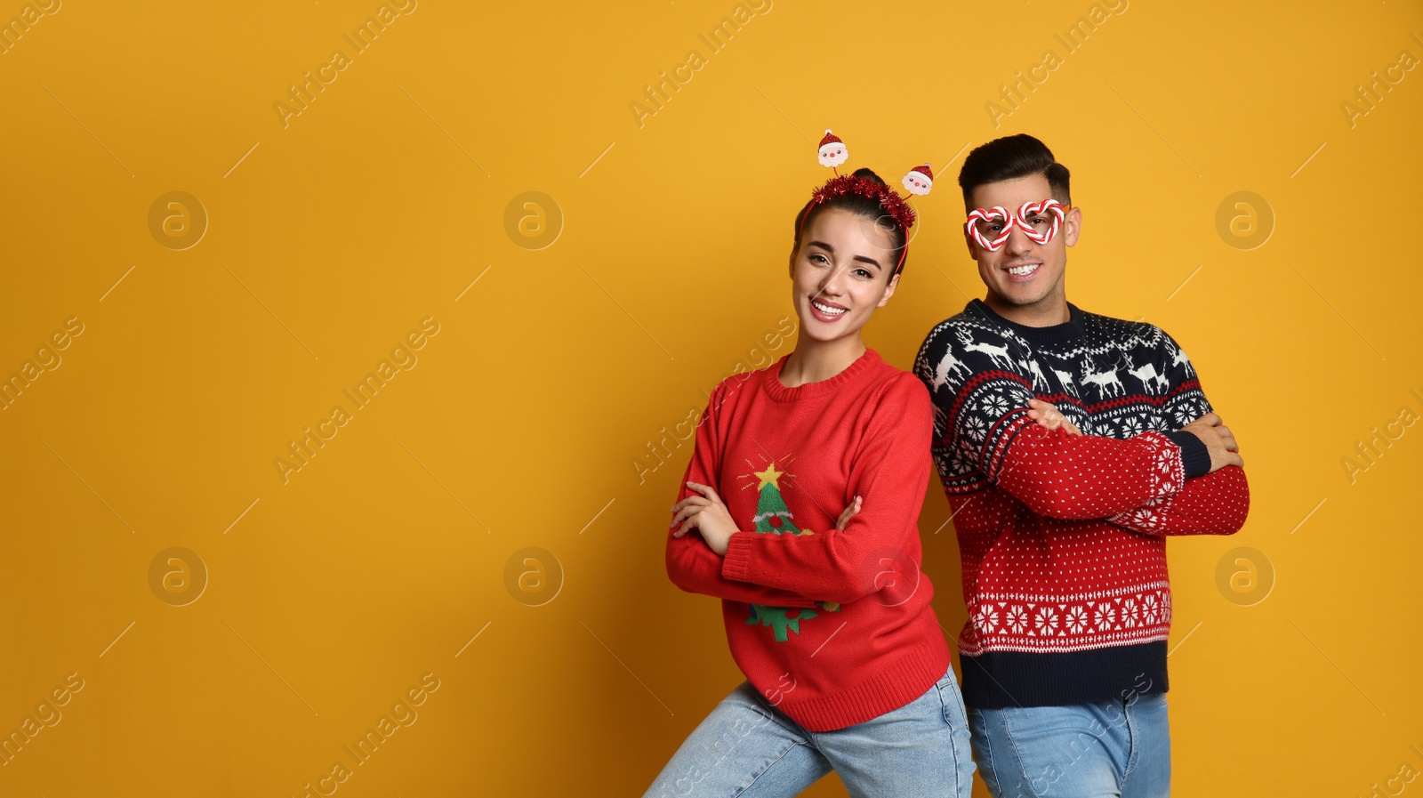 Photo of Couple in Christmas sweaters, Santa headband and party glasses on yellow background, space for text