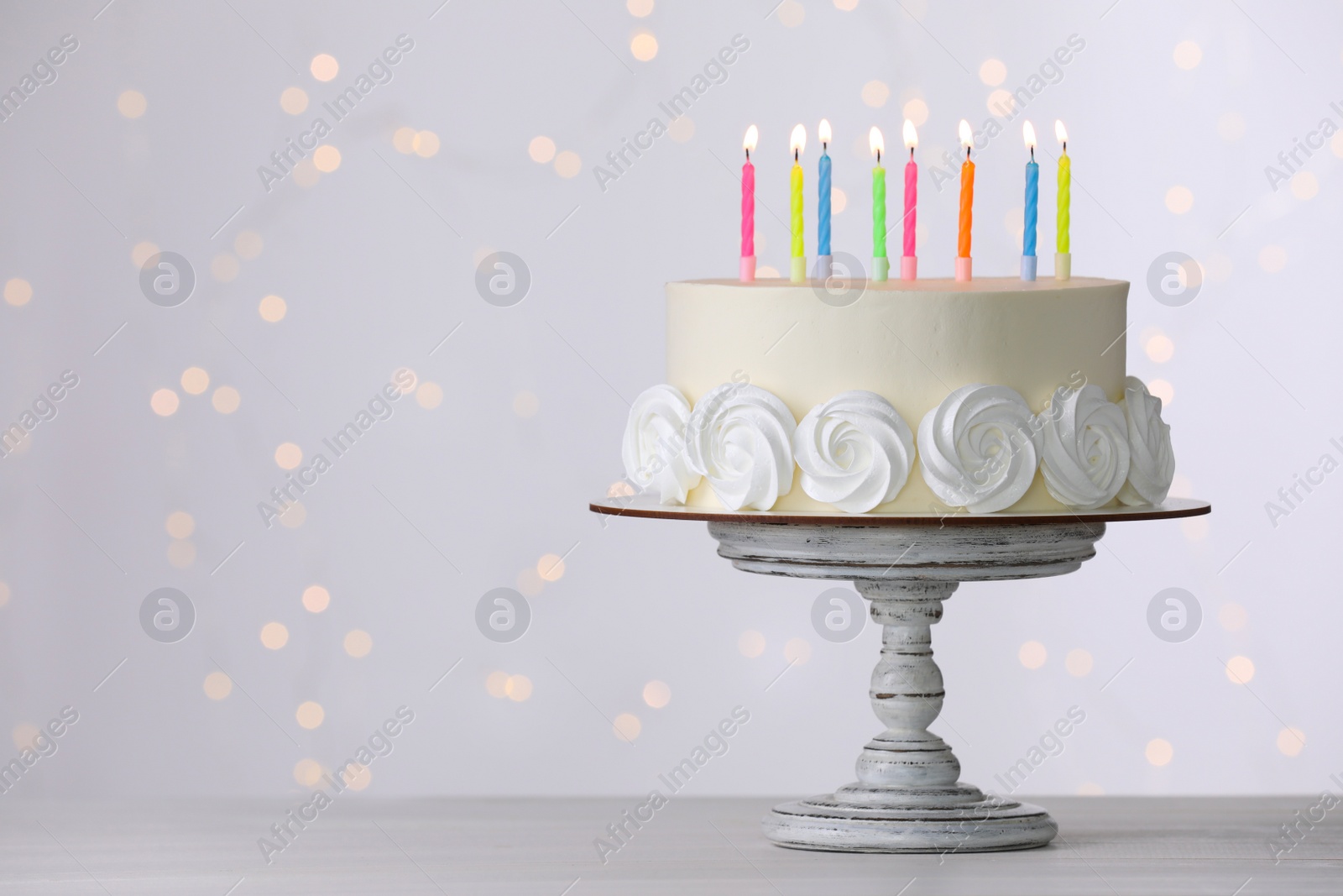 Photo of Birthday cake with burning candles on white table against blurred festive lights, space for text