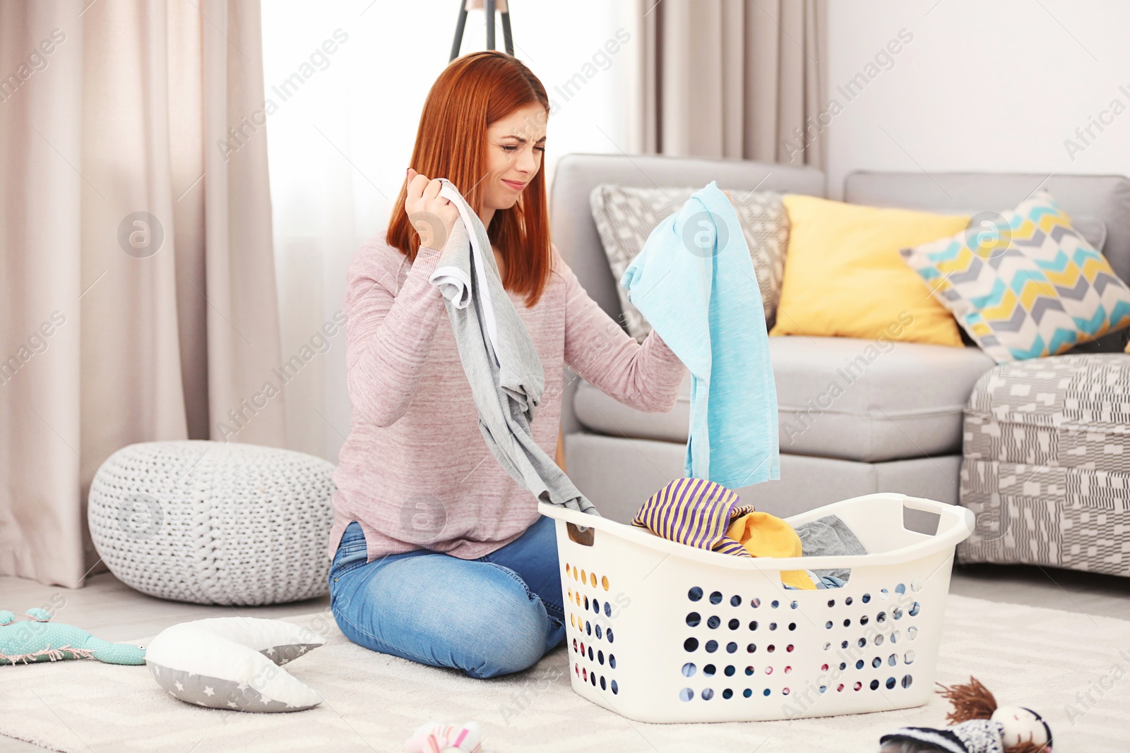 Photo of Tired housewife with basket full of clothes sitting on carpet at home