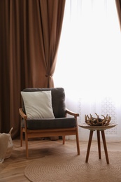 Comfortable armchair with cushion near window with stylish curtains in living room. Interior design