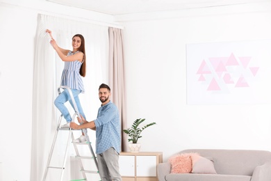 Young couple hanging window curtain in room. Space for text