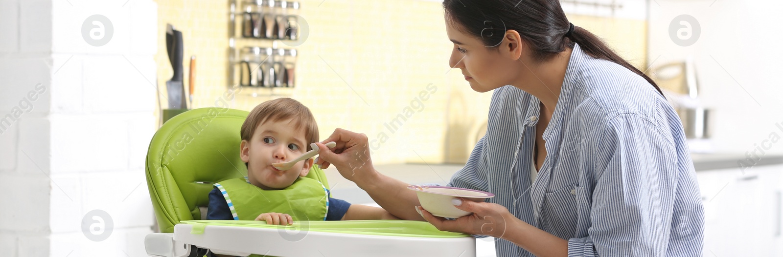 Image of Young nanny feeding cute little baby in kitchen. Banner design 