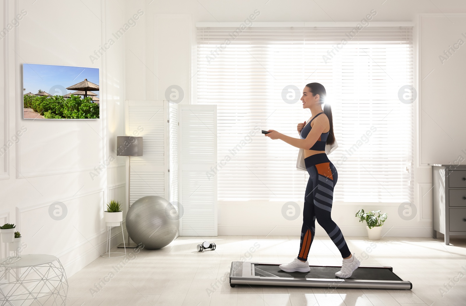 Image of Sporty woman with remote control training on walking treadmill and watching TV at home