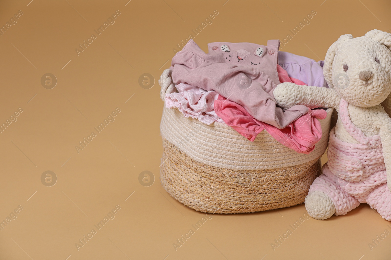 Photo of Laundry basket with baby clothes near soft toy on light brown background. Space for text
