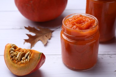 Jars of delicious pumpkin jam, fresh pumpkin and dry leaves on white wooden table, closeup
