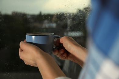 Photo of Young woman holding cup of coffee at window with rain drops, closeup