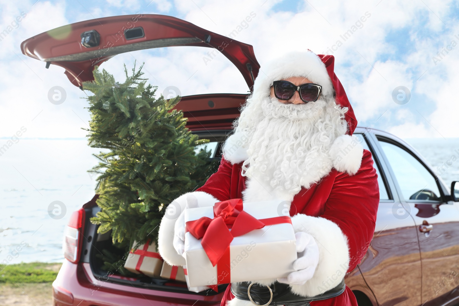 Photo of Authentic Santa Claus near red car with gift boxes and Christmas tree on beach