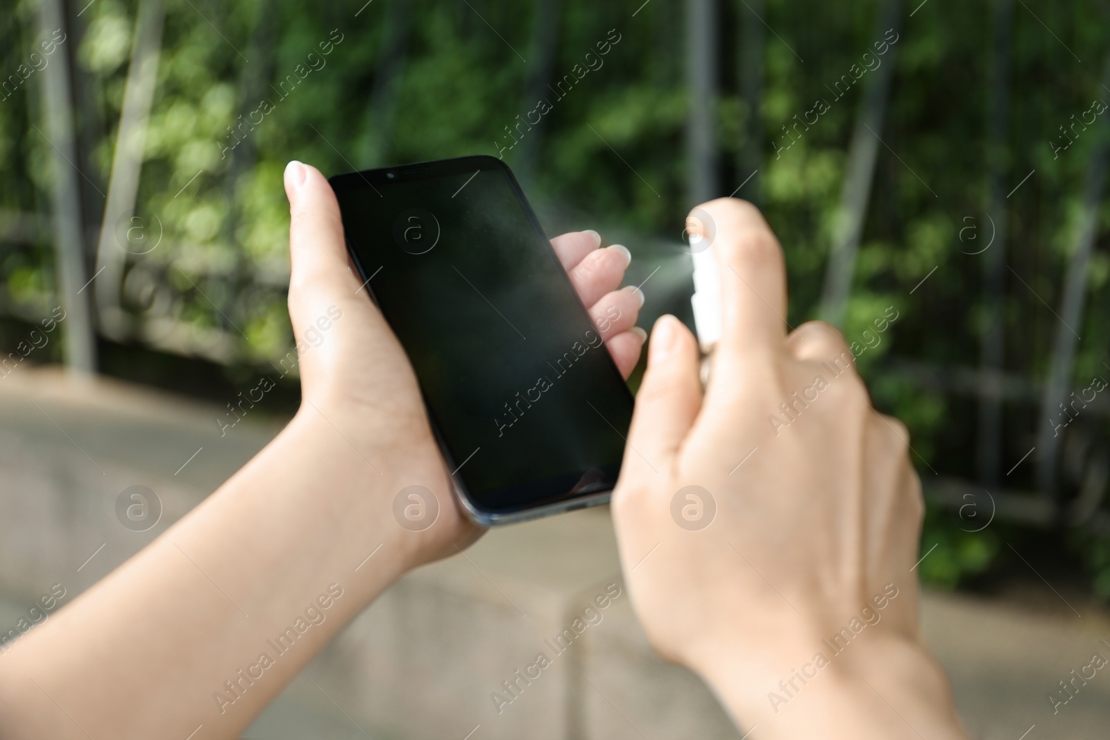 Photo of Woman spraying antiseptic onto smartphone outdoors, closeup