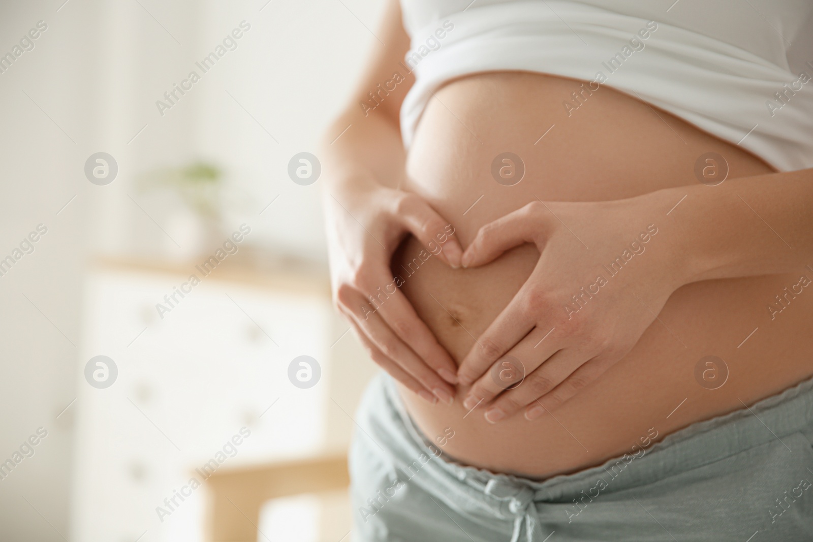 Photo of Pregnant woman making heart with her hands near belly indoors, closeup