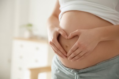 Photo of Pregnant woman making heart with her hands near belly indoors, closeup